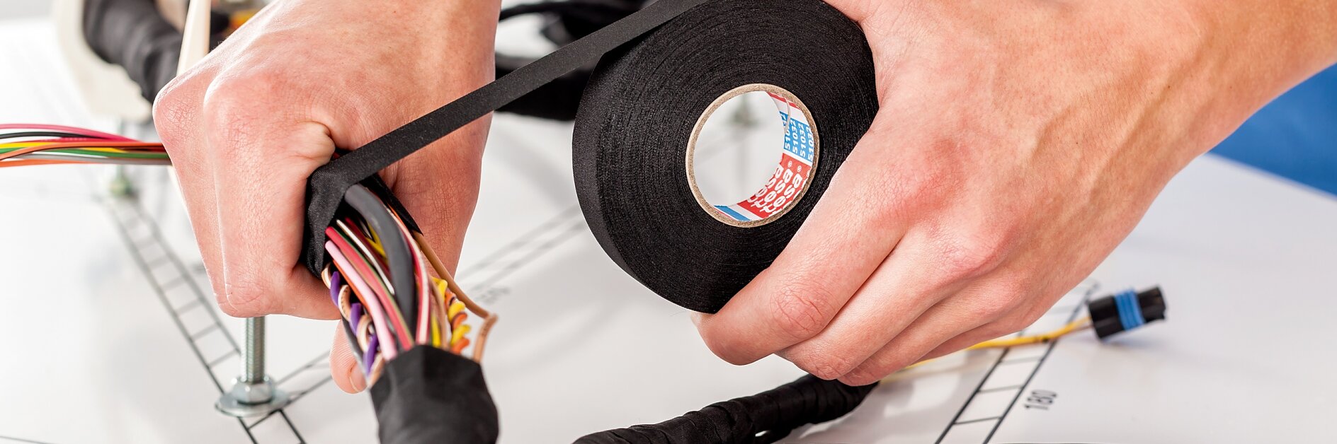 Hands wrapping black tesa tape around a bundle of colorful wires on a table. Another roll of tesa tape and more wires are visible in the background. (This text has been generated by AI)