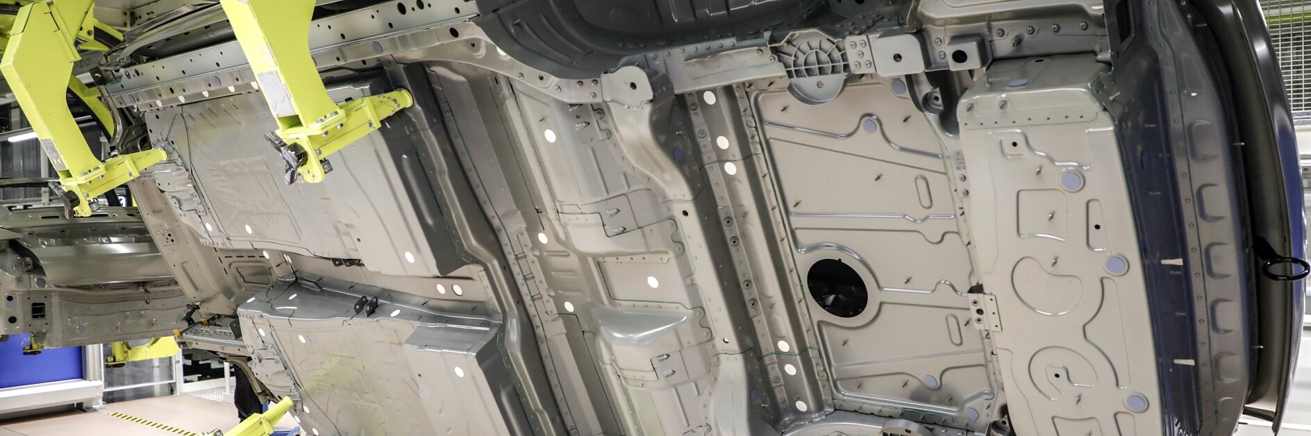 The image shows a vehicle underbody on an assembly line. The car body is held horizontally by large yellow robotic arms in a factory setting, revealing details of the chassis and structural components. Any references to tape in this context would involve tesa tape for securing or managing components efficiently during assembly. (This text has been generated by AI)