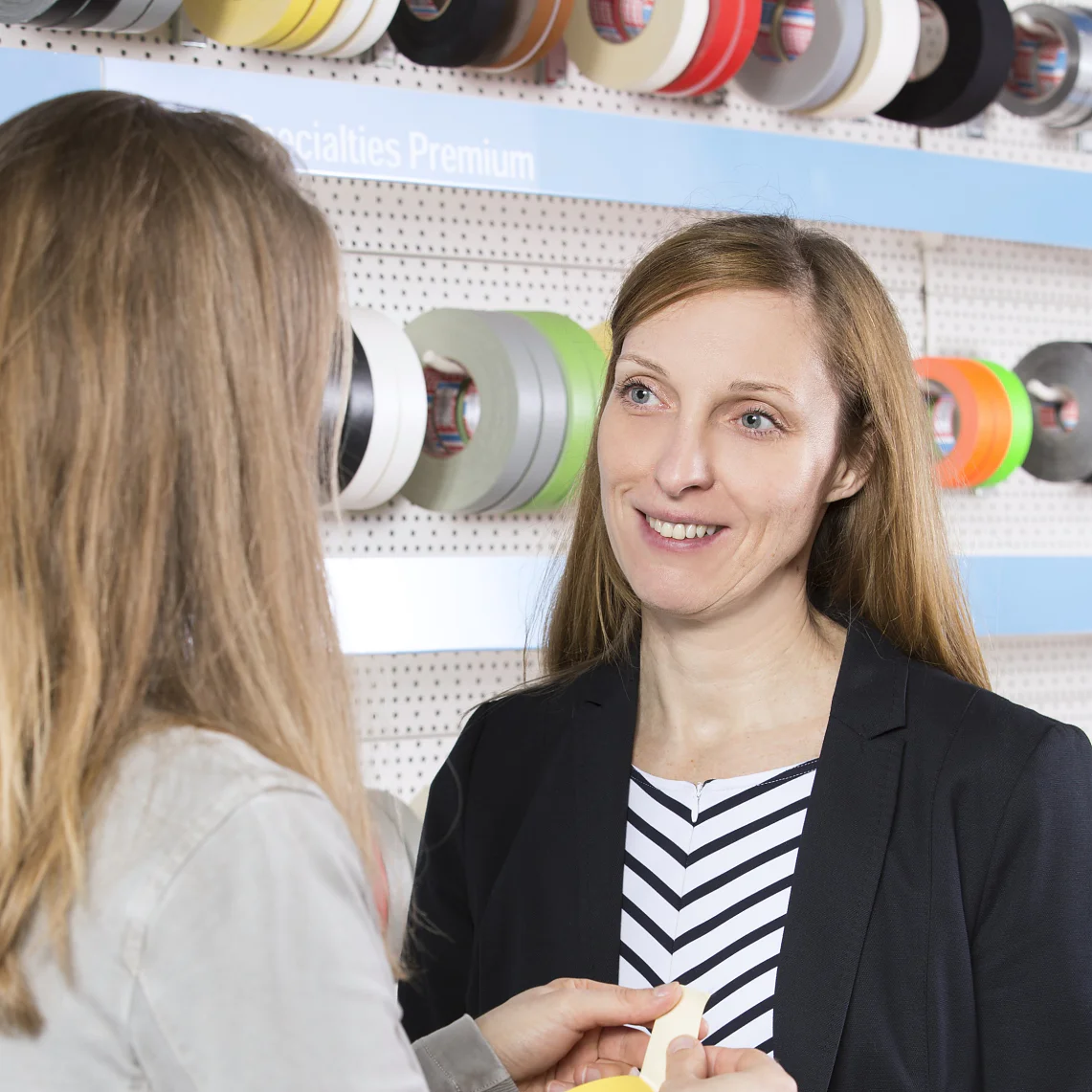 Two women are standing in front of a display of colorful tesa tape on a pegboard. One woman, in a striped shirt and blazer, is facing the camera, while the other, with long hair, is turned away, holding some samples. (This text has been generated by AI)