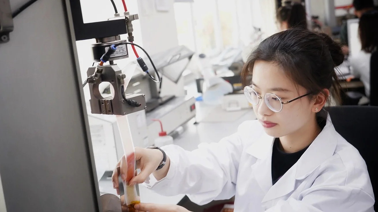 A woman in a white lab coat conducts an experiment in a laboratory. She is operating a piece of machinery and using her hands to adjust an object with tesa tape. She is focused on her work, and the lab is equipped with various scientific instruments. (This text has been generated by AI)