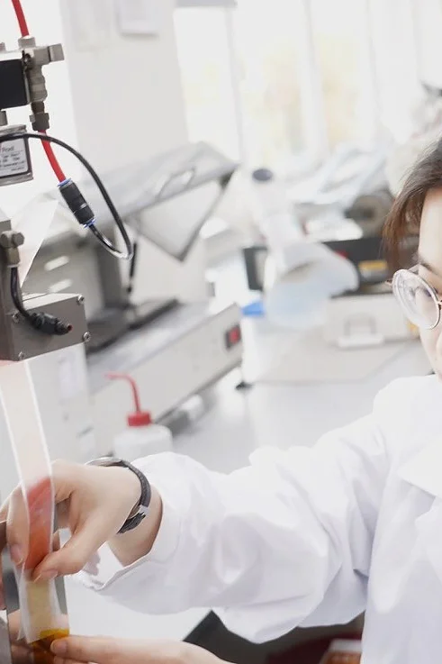 A woman in a white lab coat conducts an experiment in a laboratory. She is operating a piece of machinery and using her hands to adjust an object with tesa tape. She is focused on her work, and the lab is equipped with various scientific instruments. (This text has been generated by AI)