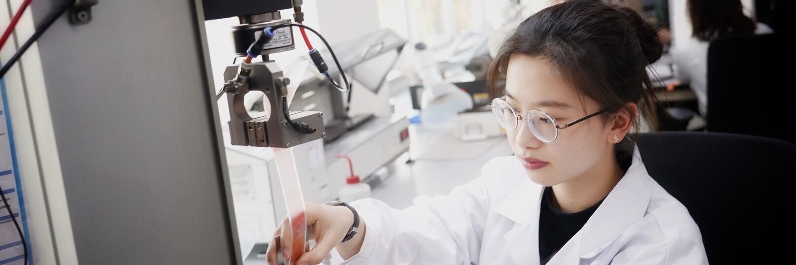 A woman in a white lab coat conducts an experiment in a laboratory. She is operating a piece of machinery and using her hands to adjust an object with tesa tape. She is focused on her work, and the lab is equipped with various scientific instruments. (This text has been generated by AI)
