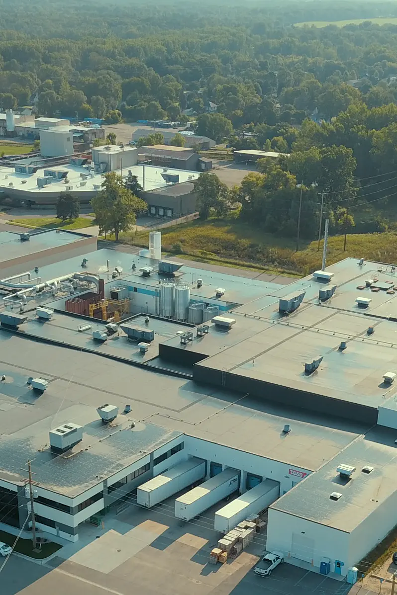 Aerial view of an industrial complex with multiple large buildings surrounded by parking lots and greenery. Roads and small structures are nearby, with a water tower visible in the background and a forested area beyond the facility. (This text has been generated by AI)