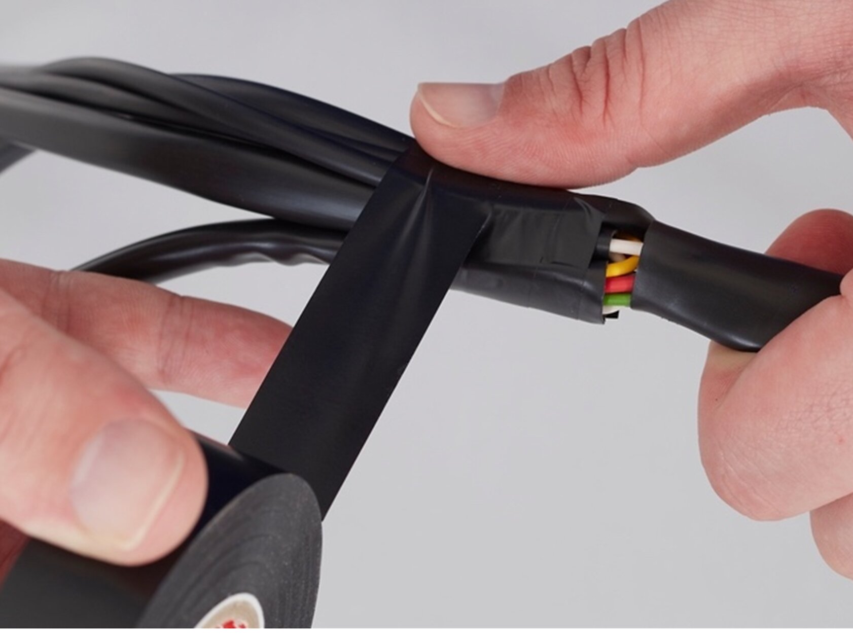 A person wraps black tesa tape around a group of colored wires. The tesa tape is being applied to secure and insulate the wires, which include green, yellow, and red colors. The background is plain white. (This text has been generated by AI)