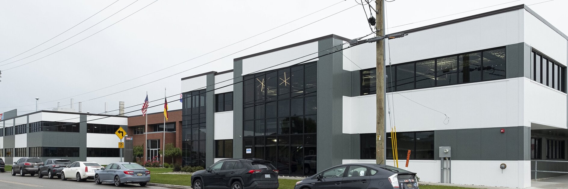 A modern office building with large glass windows and a mix of white and gray panels stands prominently. Several cars are parked along the street in front of it. Two flagpoles with flags are situated near the entrance, and an electricity pole is visible in the foreground. (This text has been generated by AI)