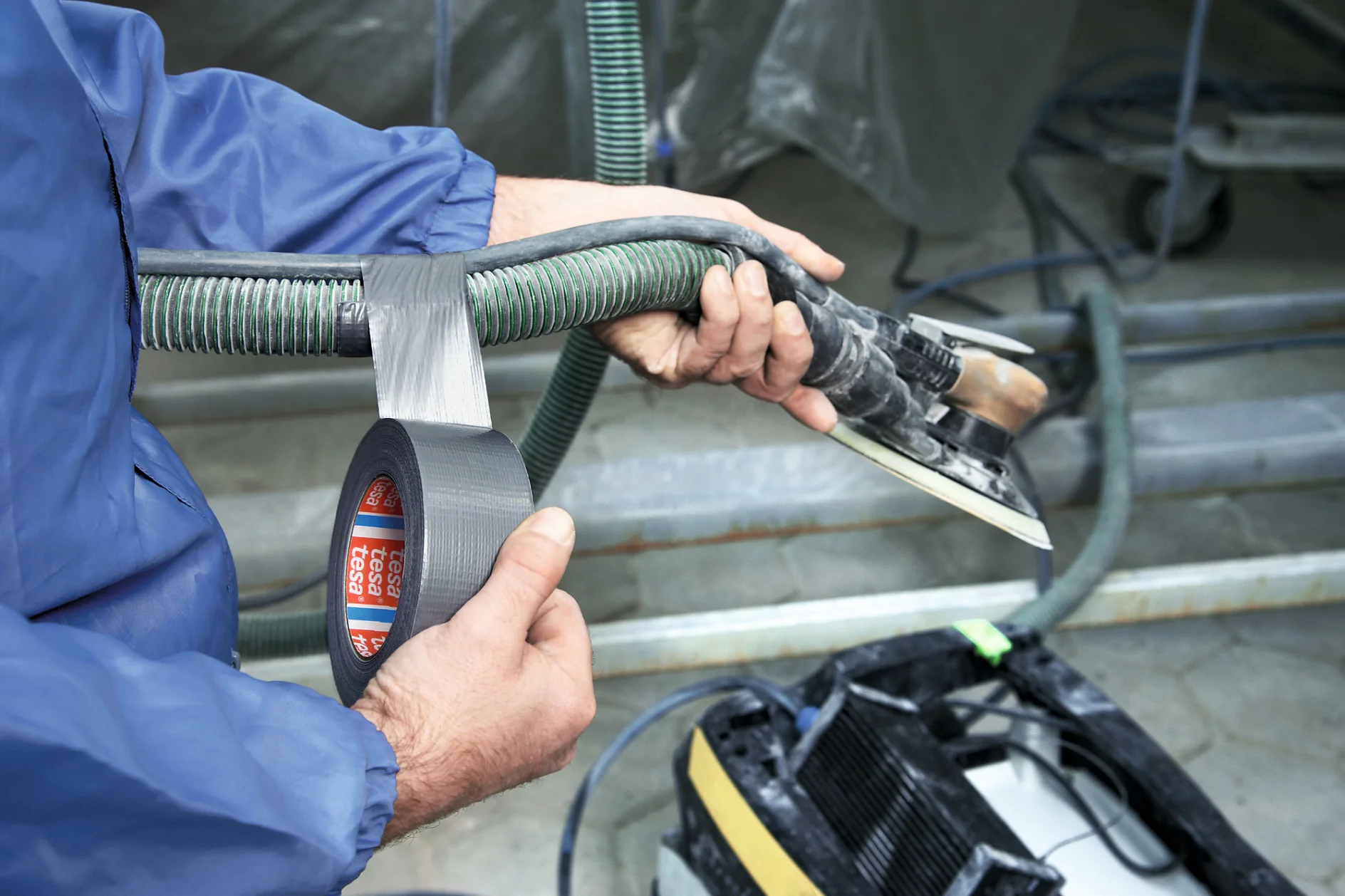 A person in a blue jacket is using silver tesa tape to secure a green ribbed hose to a metal tool with a rectangular base. The tool is connected to a piece of equipment with wheels in the background. (This text has been generated by AI)
