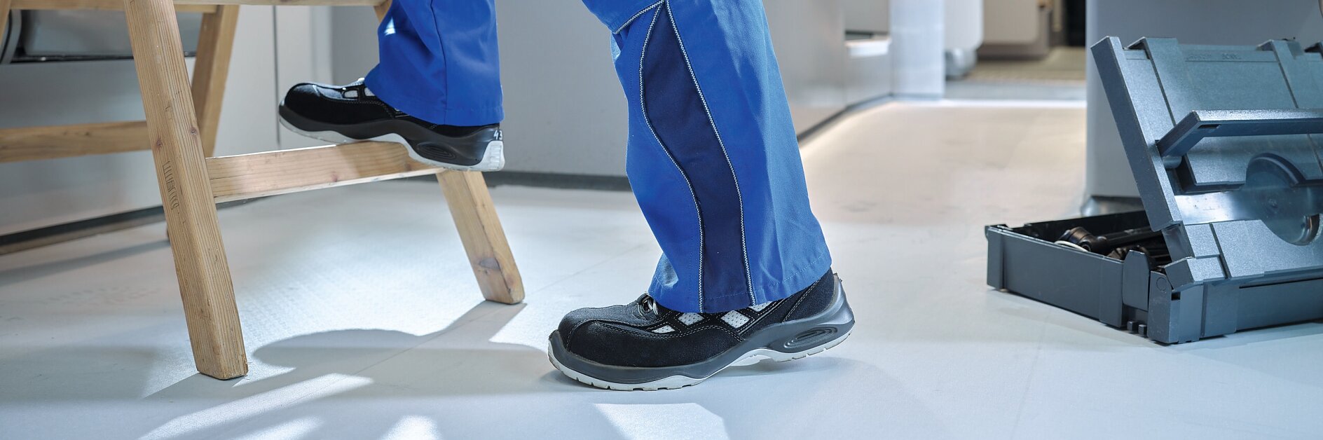 A person wearing blue pants and black shoes is ascending a wooden ladder inside a train car. The steps of the ladder are covered with anti slip tape for better grip. A toolbox with open compartments is on the floor nearby, filled with various tools. The interior of the train features red and wooden seats. (This text has been generated by AI)