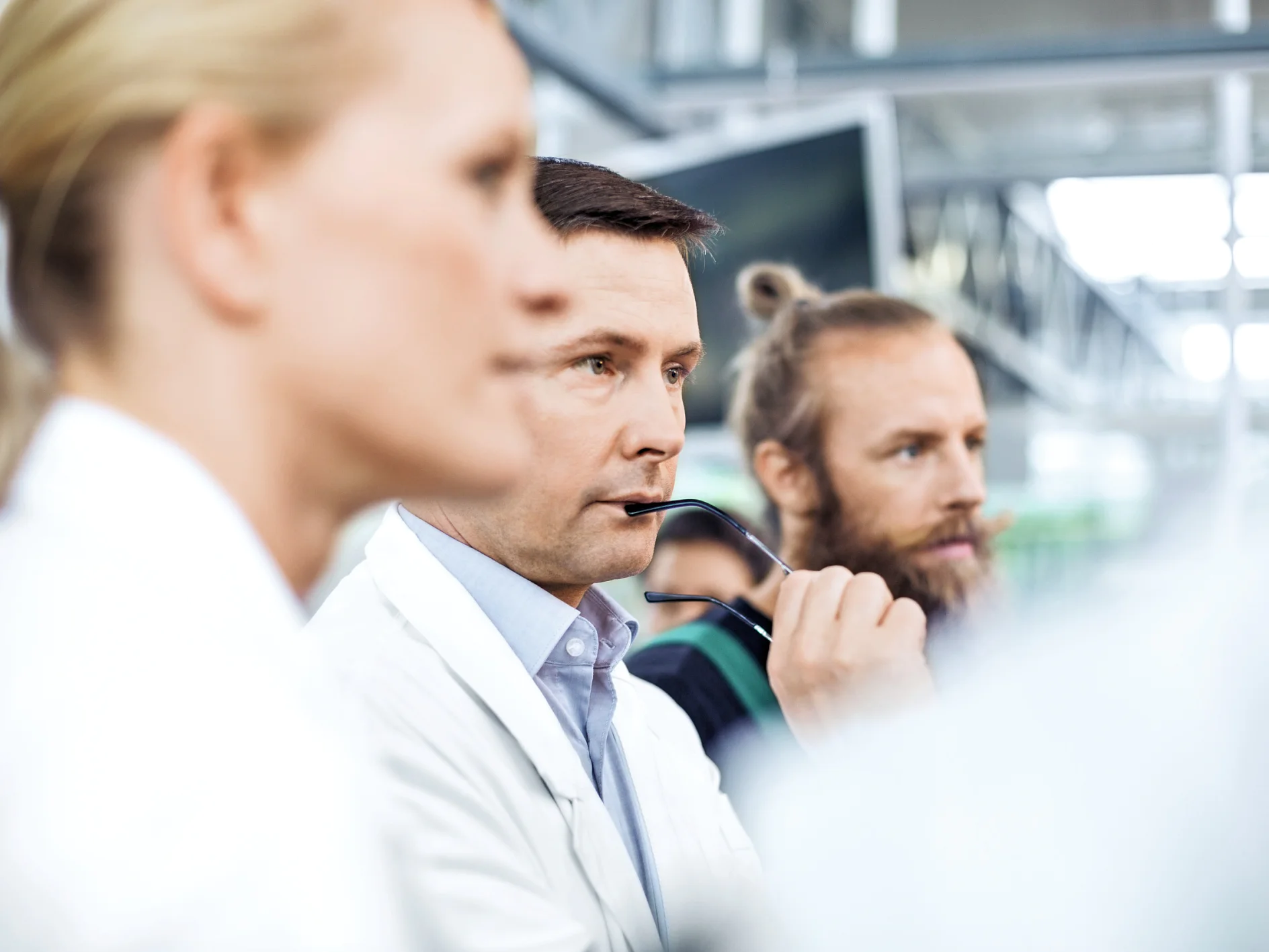 Three people indoors listen intently; the man in the center holds a pen. All wear business or lab attire in a modern workspace. (This text has been generated by AI)