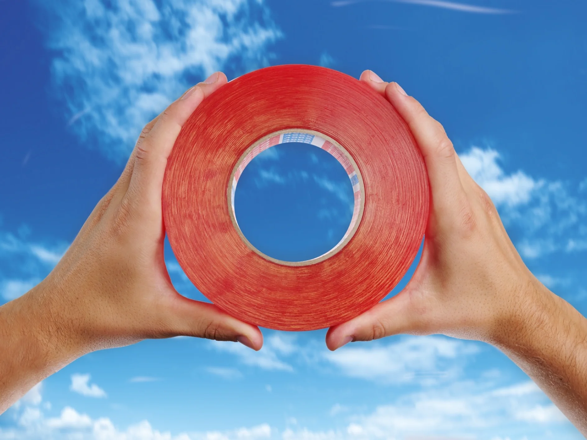 tesa-ITC Mounting_Samevalue_Inset-1 Two hands hold a large roll of red tesa tape against a backdrop of a blue sky with scattered clouds. The tesa tape roll forms a circular frame through which the sky is visible. (This text has been generated by AI)