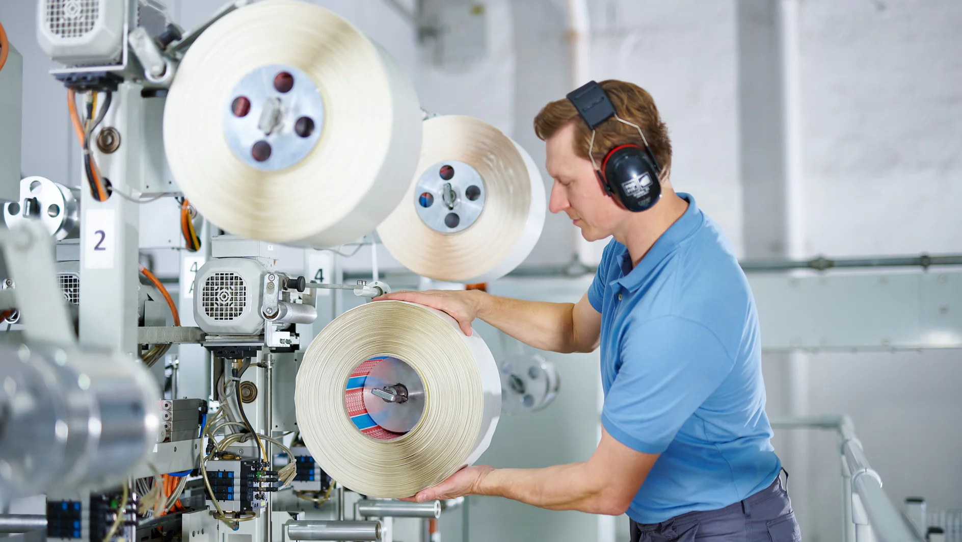 A man wearing a blue shirt and protective earmuffs operates machinery in an industrial setting. He is handling a large spool of tesa tape. There are multiple spools of tesa tape on the machine, and various mechanical components are visible. (This text has been generated by AI)