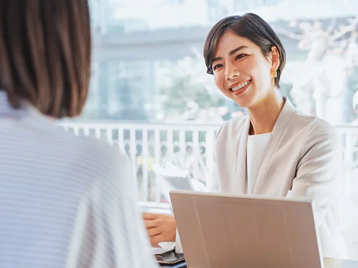 A woman in business attire sits at a desk with a laptop, smiling and talking to another person. tesa tape is on the desk. (This text has been generated by AI)