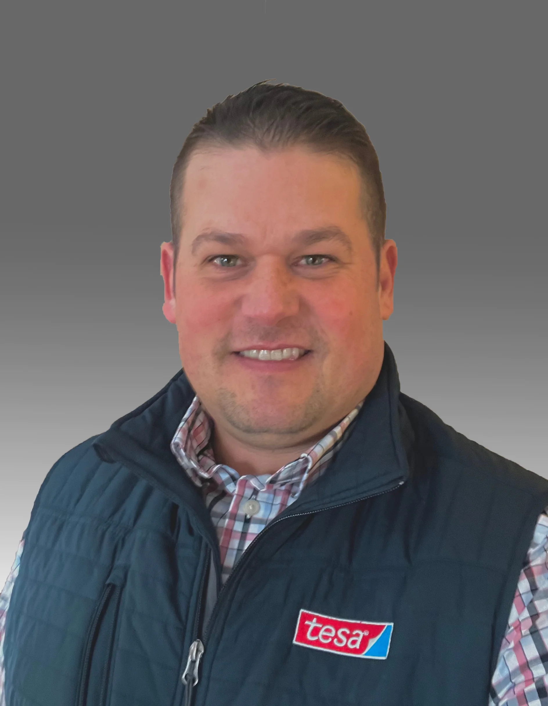 A person with short, dark hair is wearing a dark blue quilted vest with a red and white logo on the chest. They have a plaid shirt underneath. The background is a gray gradient. (This text has been generated by AI)