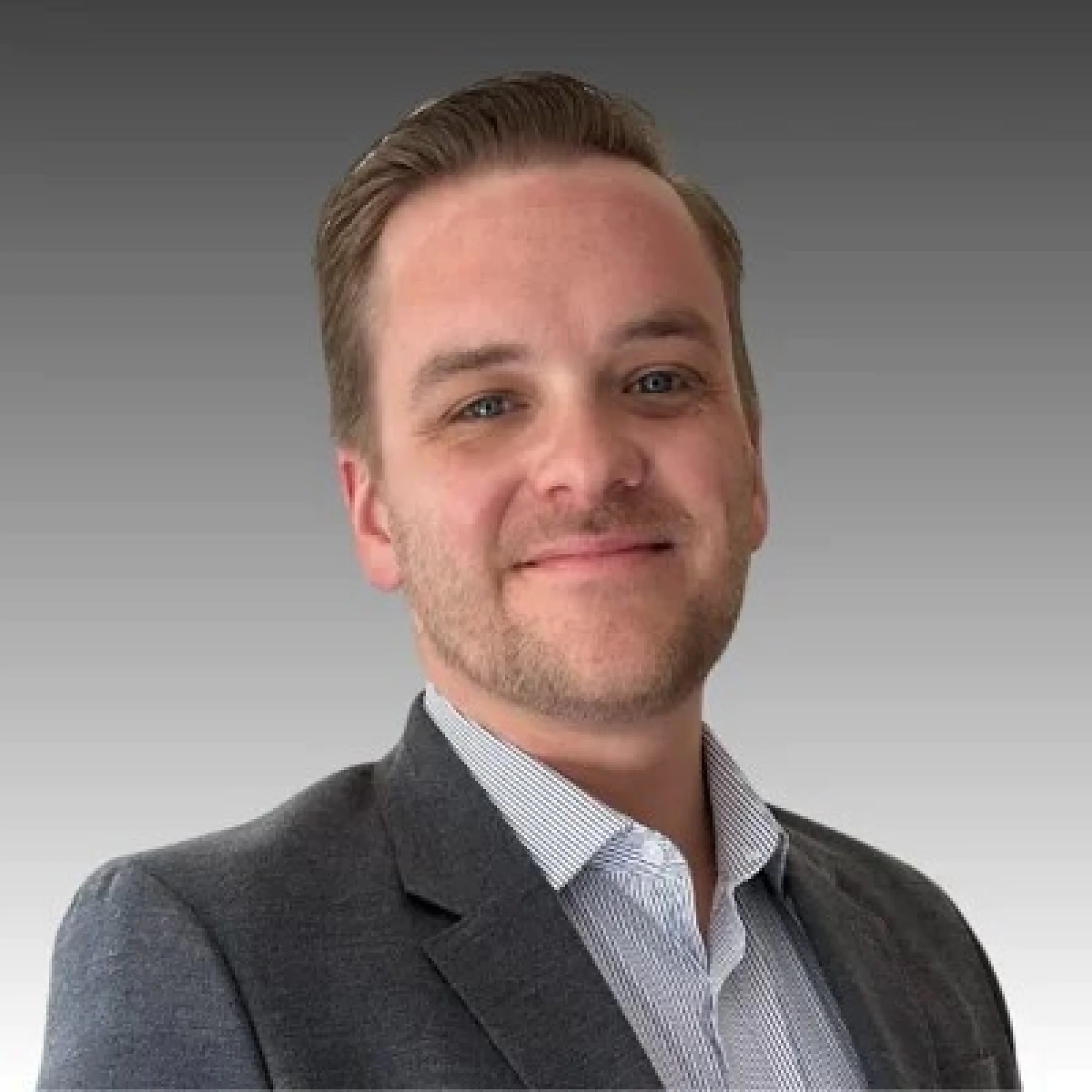 A person with short hair is smiling slightly, wearing a gray suit jacket over a striped shirt. The background is a gradient from gray to lighter gray. (This text has been generated by AI)