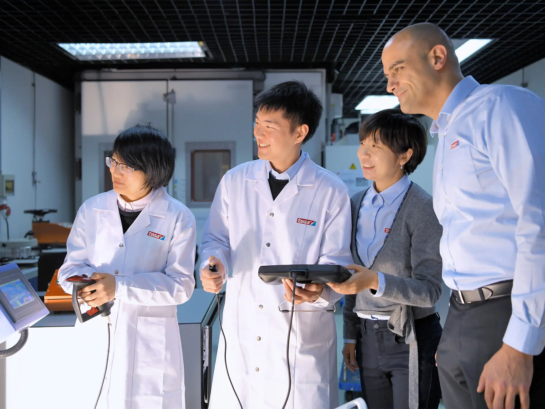 four professionals in lab coats and business attire discuss data on a tablet in a modern indoor lab setting with equipment around tesa (This text has been generated by AI)