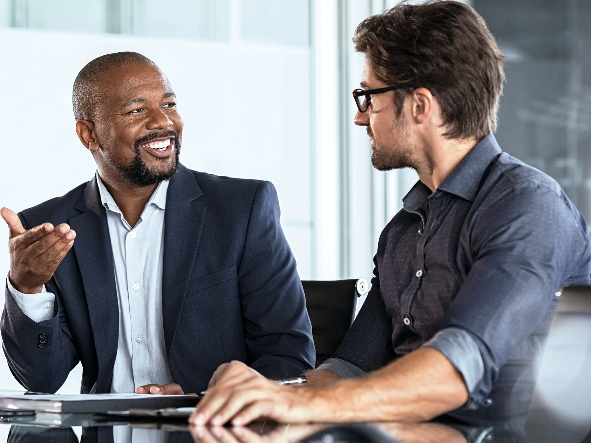 Two men in business attire engaged in discussion at an office desk with laptop and documents, indoor professional setting. (This text has been generated by AI)