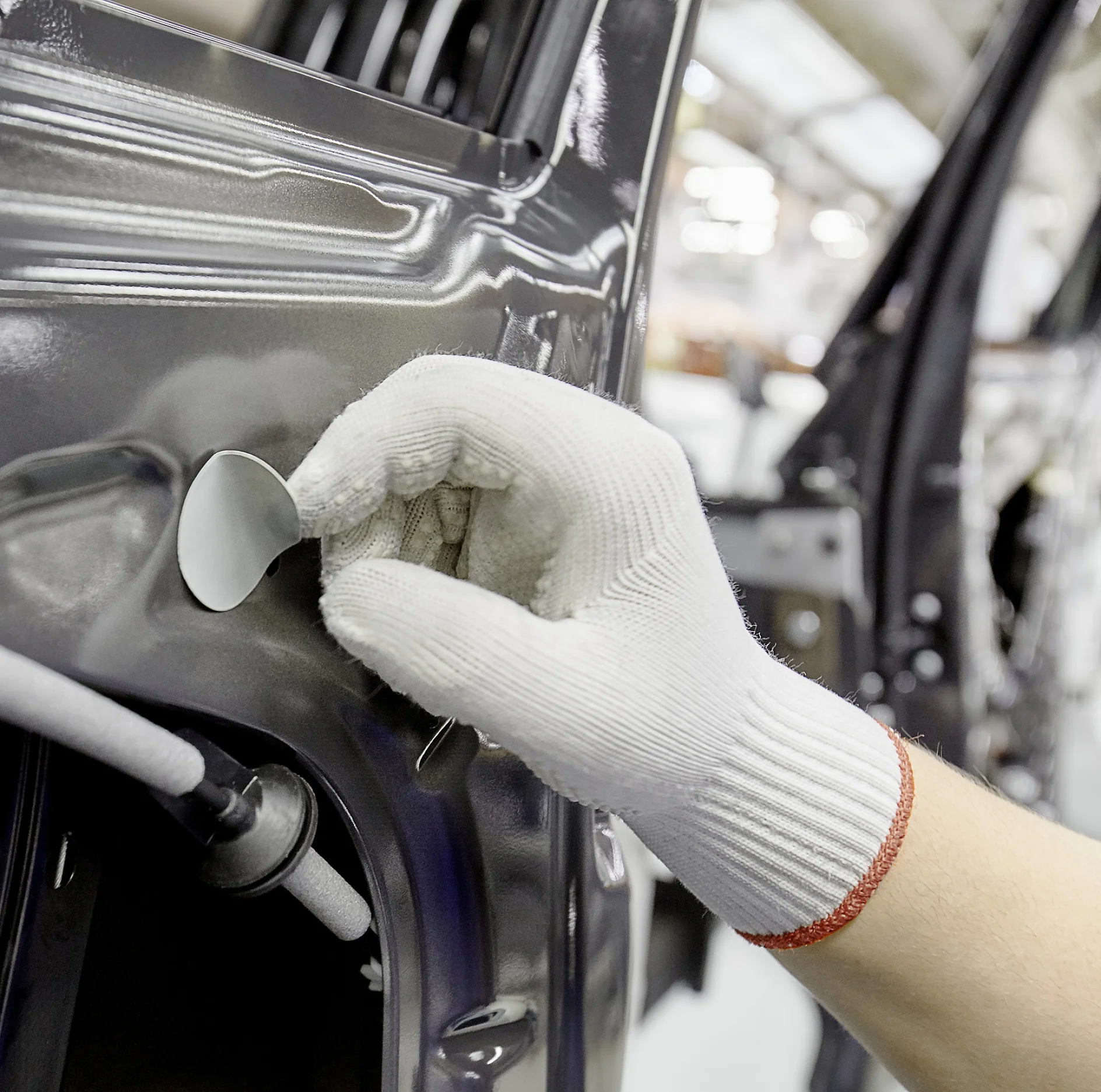 A person wearing a white work glove is applying a circular tesa tape adhesive patch to the inside of a metallic car door in an automotive assembly or repair setting. (This text has been generated by AI)