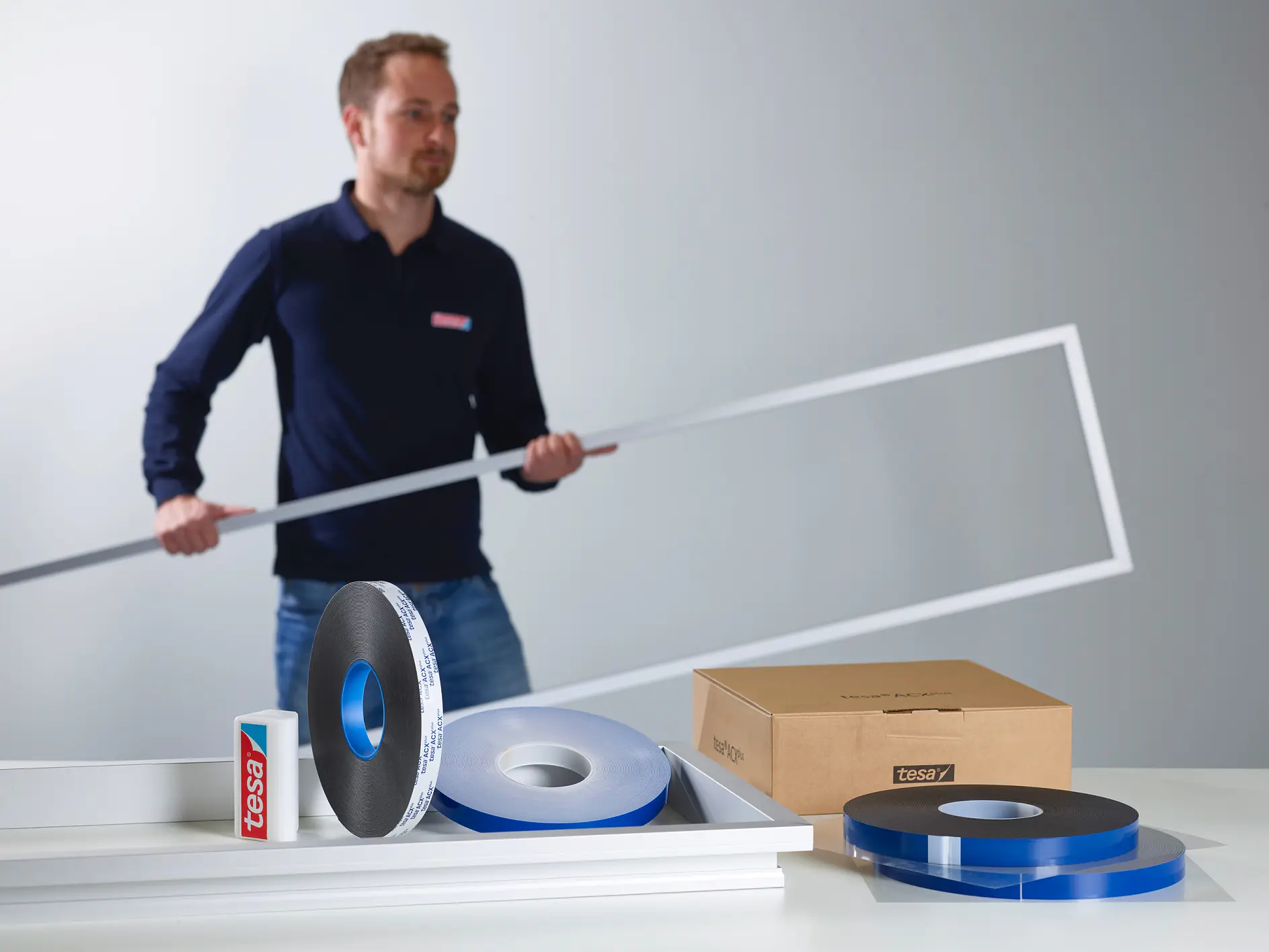 man holding a white rectangular frame standing behind a table with rolls of tape, a cardboard box, and a tesa branded container in a studio setting