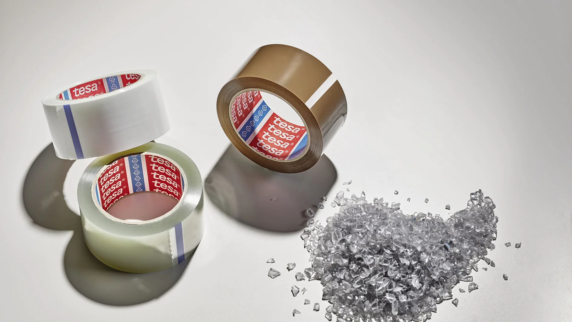 Three rolls of packing tesa tape, two clear and one brown, are arranged on a light surface. Beside them is a small pile of clear plastic granules. Shadows extend from the objects. (This text has been generated by AI)