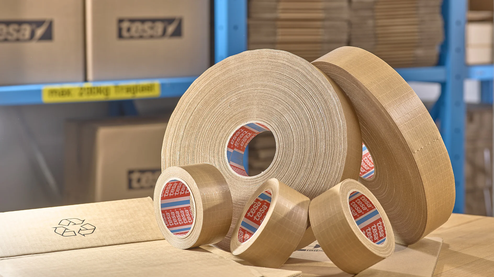 Several rolls of brown packing tesa tape in different sizes are displayed on a table. In the background, there are stacked cardboard boxes and packages on blue shelves. A cardboard box with a recycling symbol is in the foreground. (This text has been generated by AI)