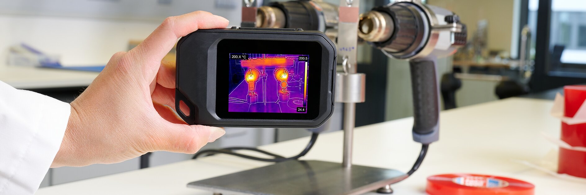 A person holds a handheld thermal imaging camera displaying heat signatures while standing in a laboratory with scientific equipment and tools, including tesa tape, visible on the counter. (This text has been generated by AI)