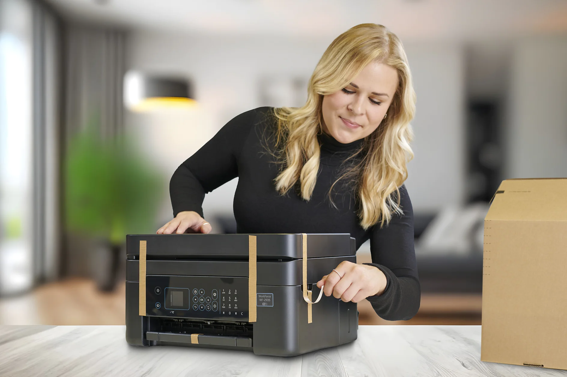 A woman with blonde hair in a black top removes tesa tape from a black printer on a white table; box right, blurred living room behind. (This text has been generated by AI)