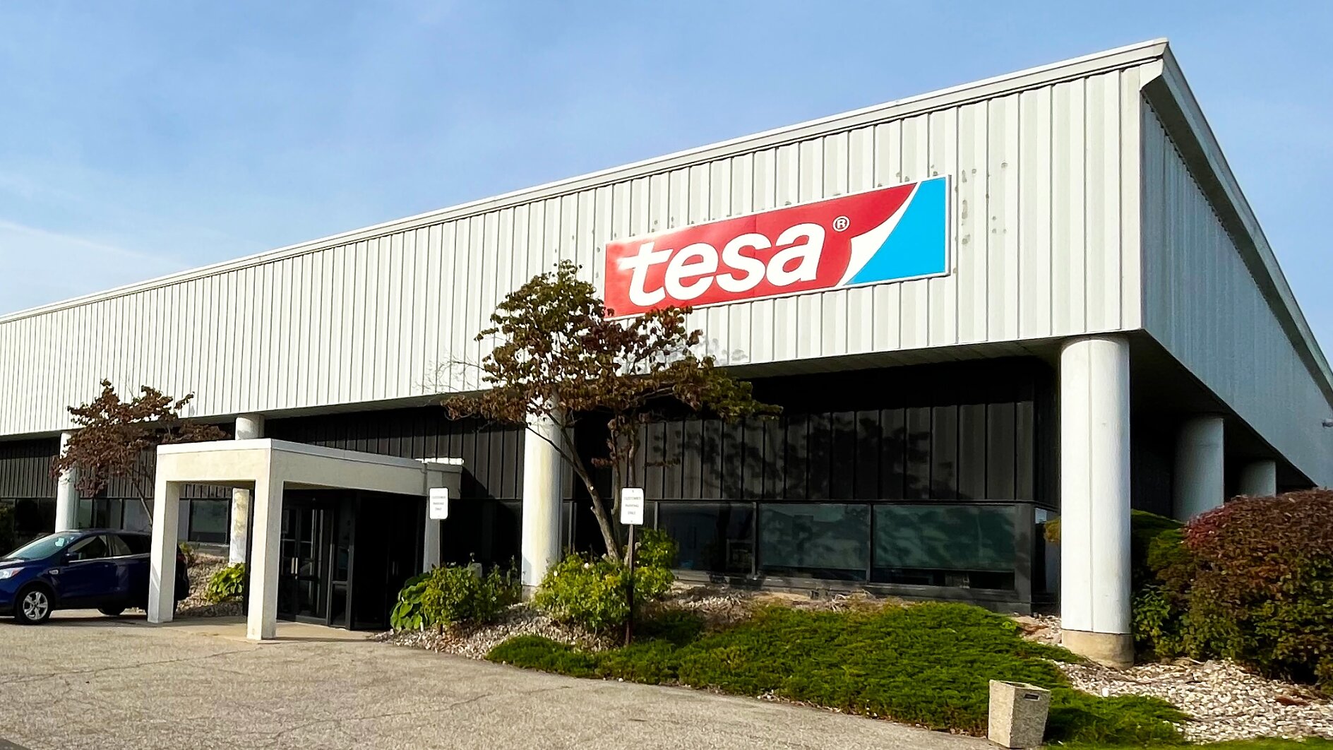 A large white industrial building features a red and blue tesa tape logo above the entrance, with small trees and a blue car in front. (This text has been generated by AI)