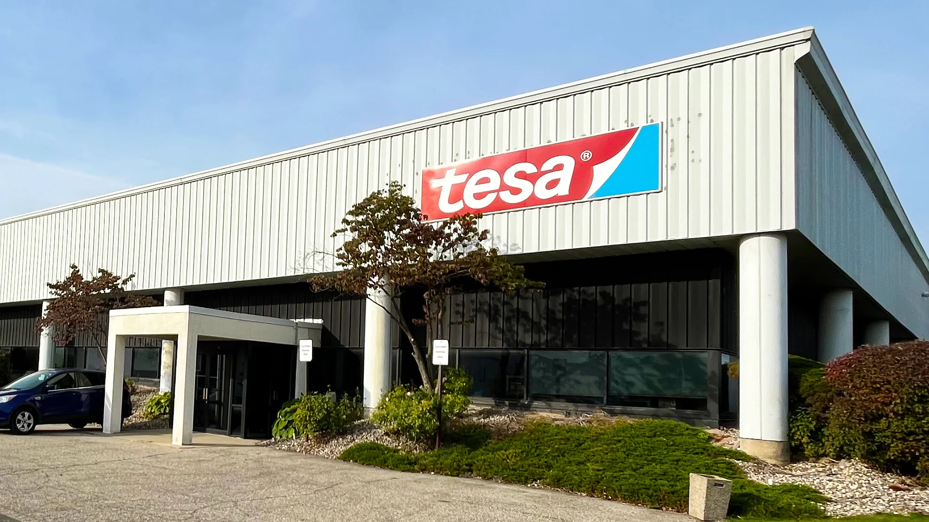 A large white industrial building features a red and blue tesa tape logo above the entrance, with small trees and a blue car in front. (This text has been generated by AI)