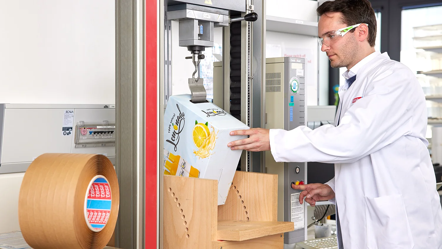 A man in a white lab coat and safety glasses operates a packaging tester near a roll of tesa tape and other lab equipment. (This text has been generated by AI)