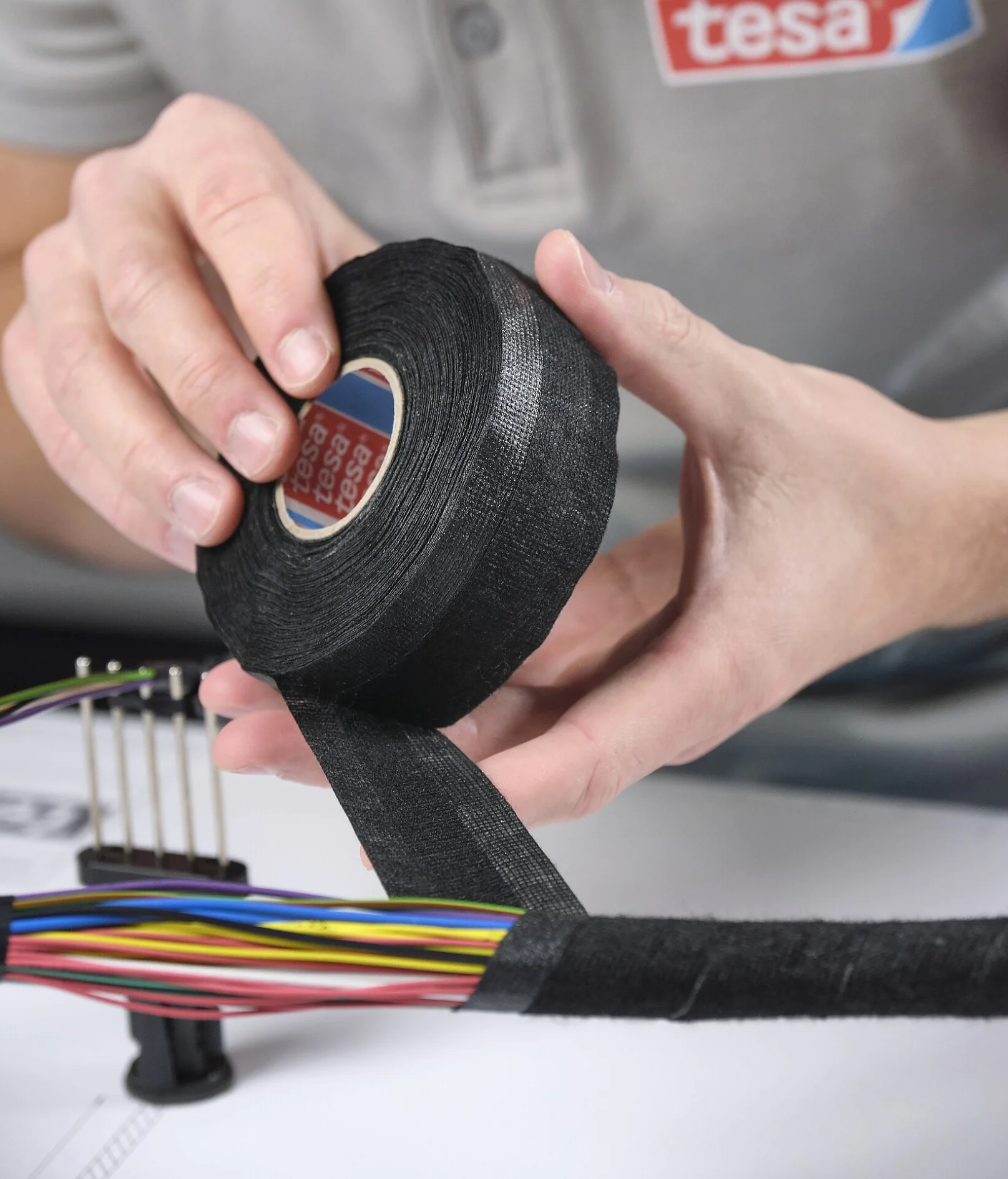 A person in a grey polo shirt wraps black tesa tape around a bundle of multicolored wires, securing and organizing them on a work surface. (This text has been generated by AI)