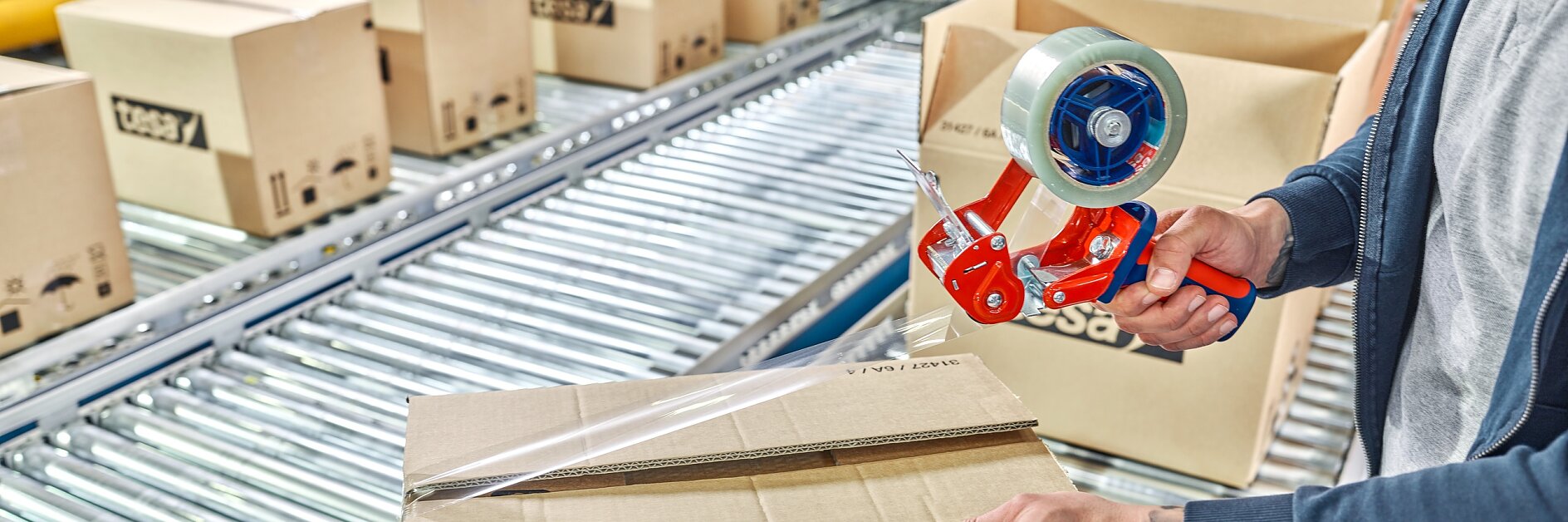 A person seals a tesa-labeled cardboard box with tesa tape using a handheld dispenser on a conveyor belt in a warehouse. (This text has been generated by AI)