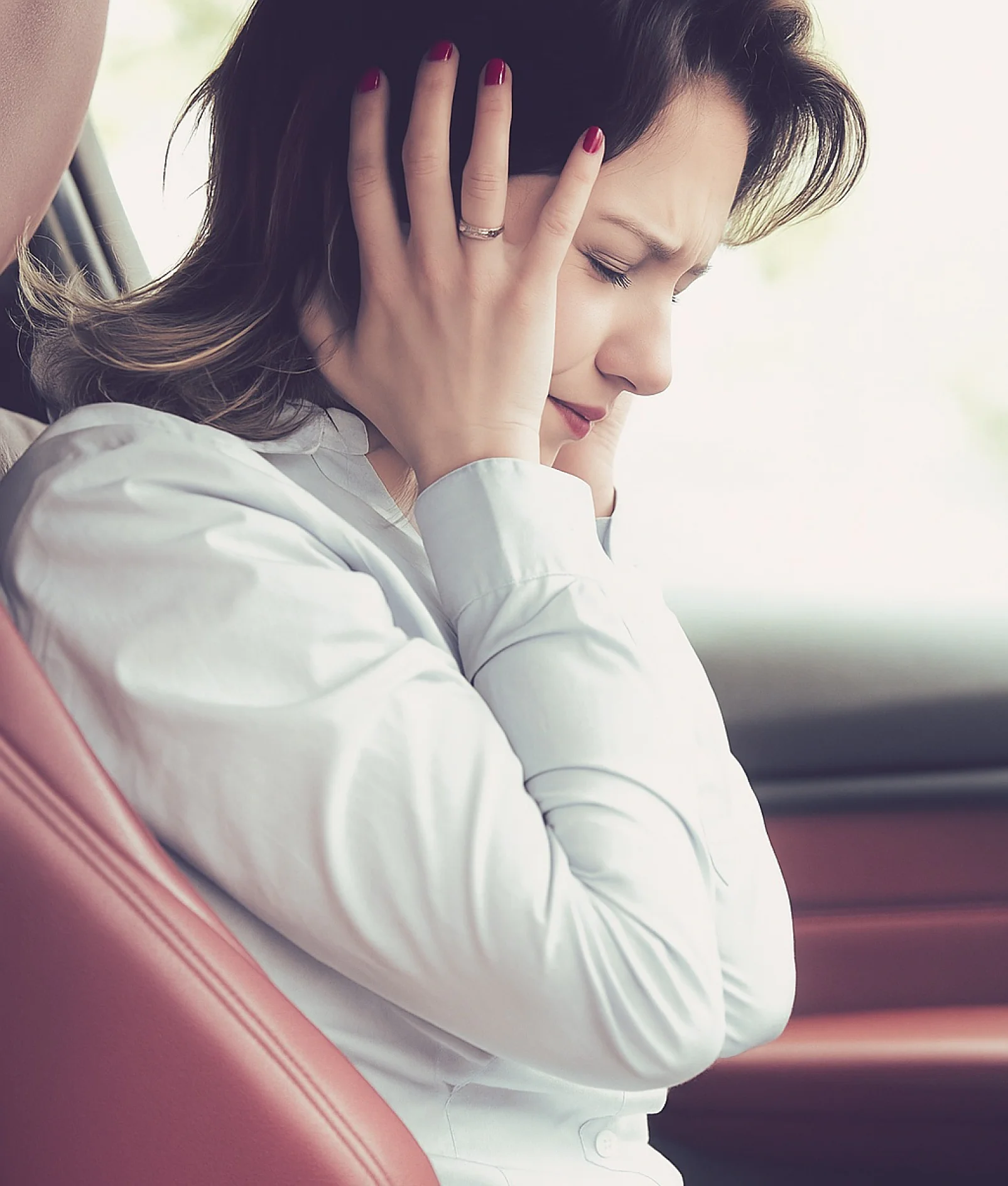 A woman sitting in a car covers her ears with both hands and closes her eyes, appearing distressed or overwhelmed. She is wearing a light blue blouse and has dark hair with lighter ends. There is visible tesa tape in the scene. (This text has been generated by AI)