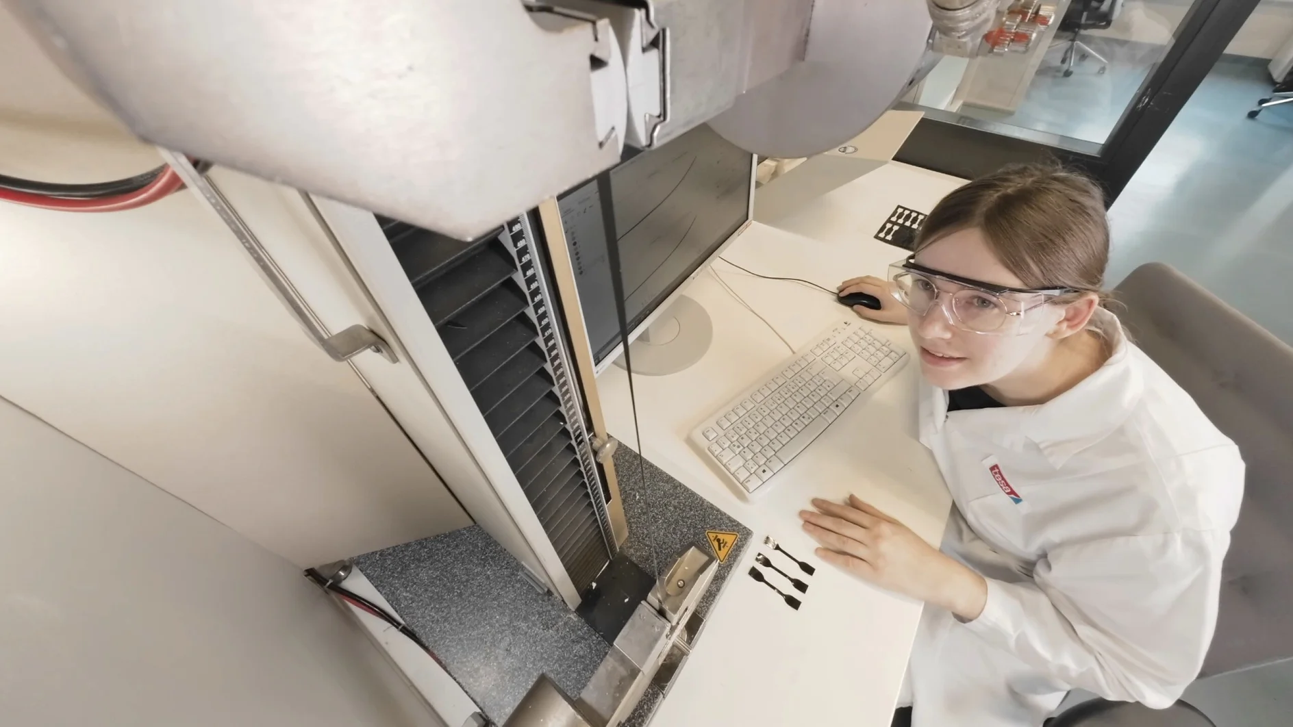 A person in safety glasses and a lab coat sits at a desk, using a computer and large equipment, with tesa tape visible nearby. (This text has been generated by AI)