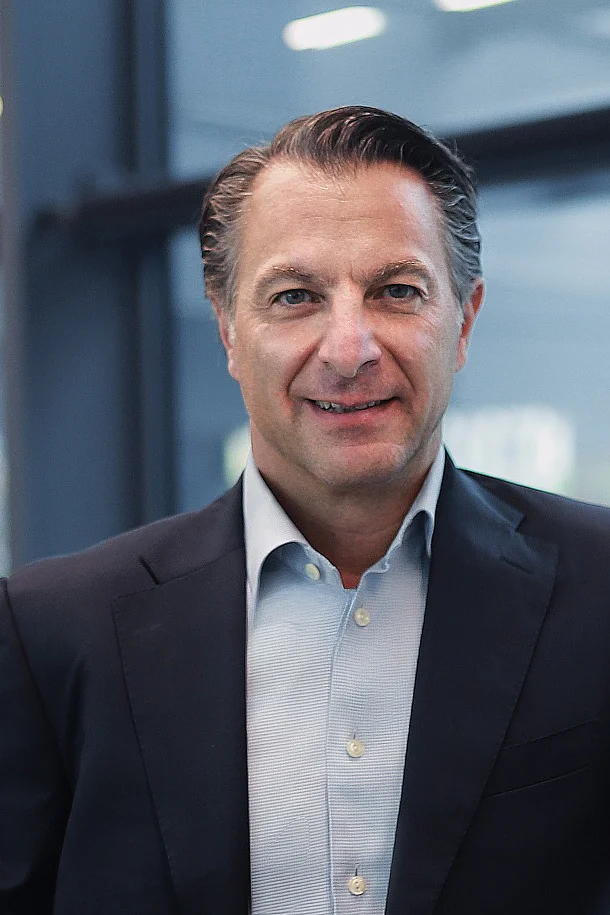 A man in a dark suit and light shirt stands by a large window indoors, one arm on a wooden railing, with glass panels and greenery outside. (This text has been generated by AI)