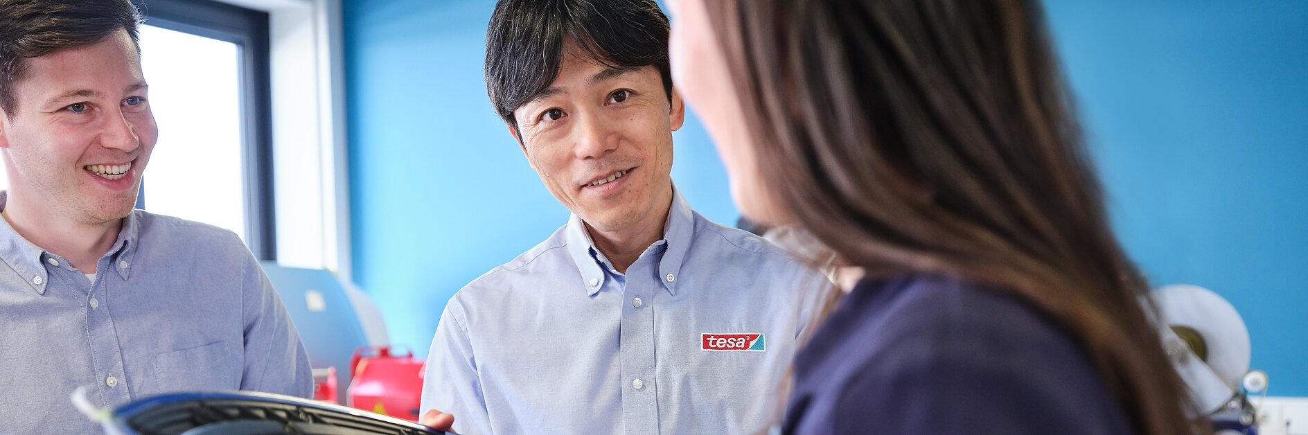 Three people are standing inside an office with blue walls. They are engaged in conversation. The person in the middle is holding a curved object, possibly related to an industrial or engineering context, and its wrapped in tesa tape. They are wearing casual business attire.