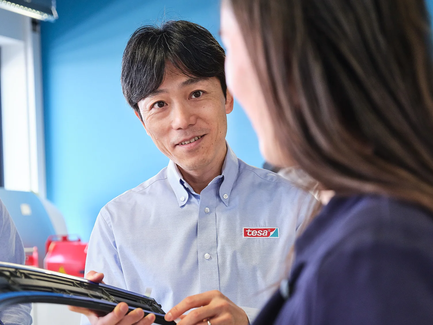 Three people are standing inside an office with blue walls. They are engaged in conversation. The person in the middle is holding a curved object, possibly related to an industrial or engineering context, and its wrapped in tesa tape. They are wearing casual business attire. (This text has been generated by AI)