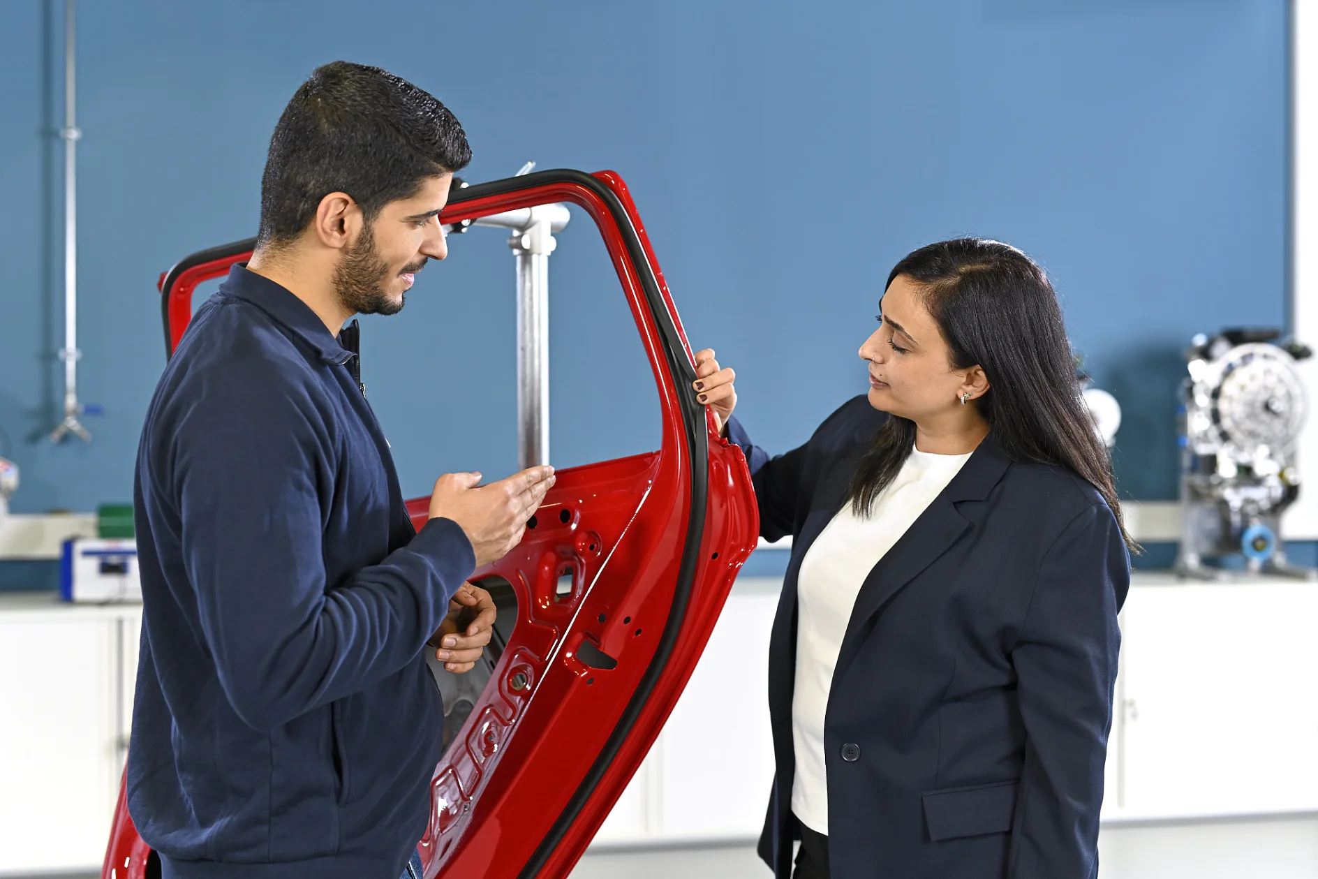 Two people by a red car door discuss; one gestures as the other listens and holds the door. Car parts and a blue wall are seen.