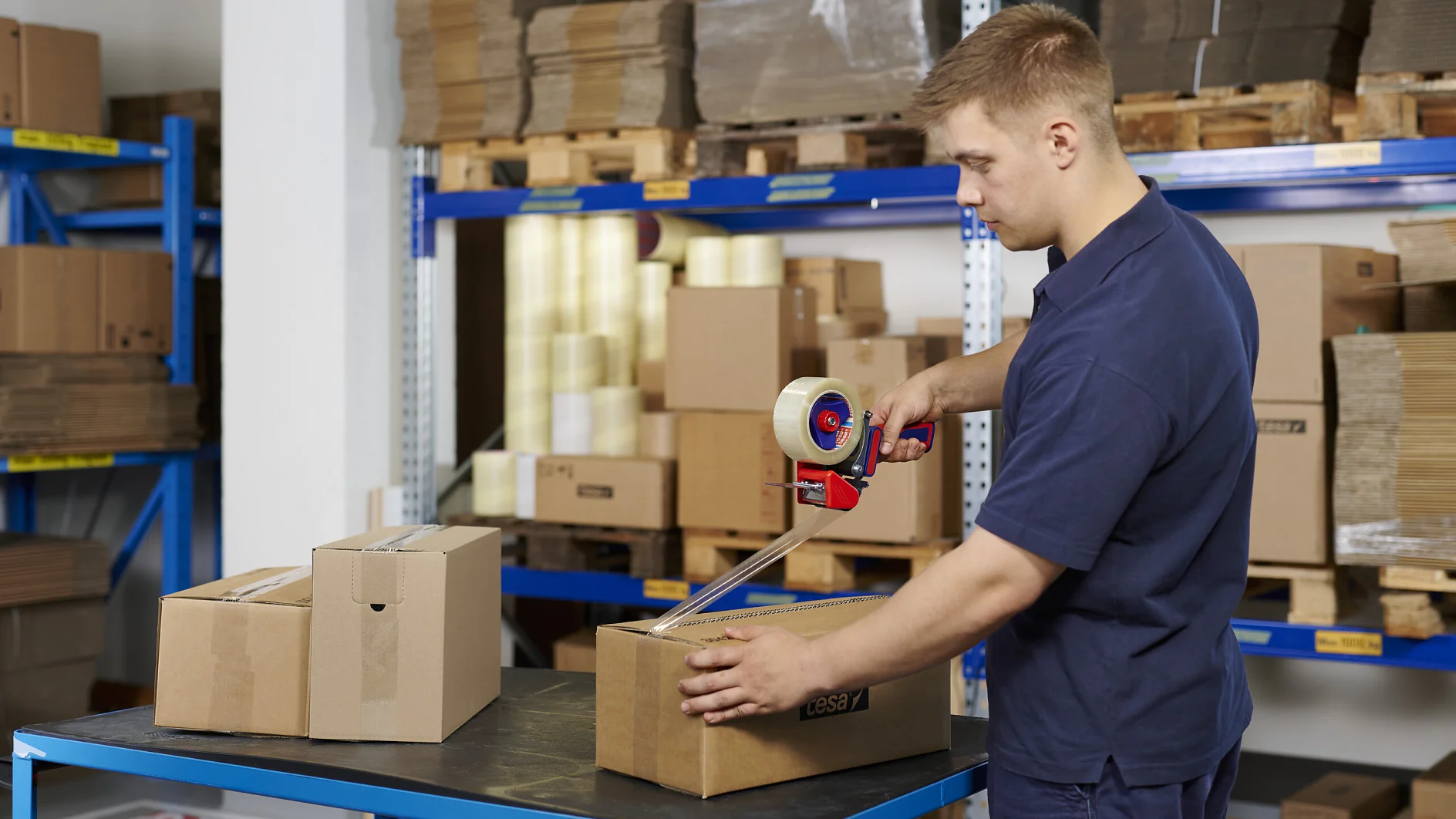 man in navy blue uniform sealing cardboard boxes with packing tape in a warehouse with shelves of stacked boxes and packaging materials (This text has been generated by AI)