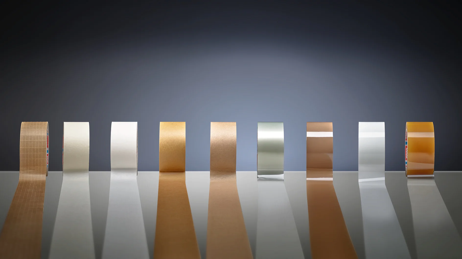 tesa adhesive tape rolls in various colors and materials lined up on a reflective surface against a dark gradient background (This text has been generated by AI)