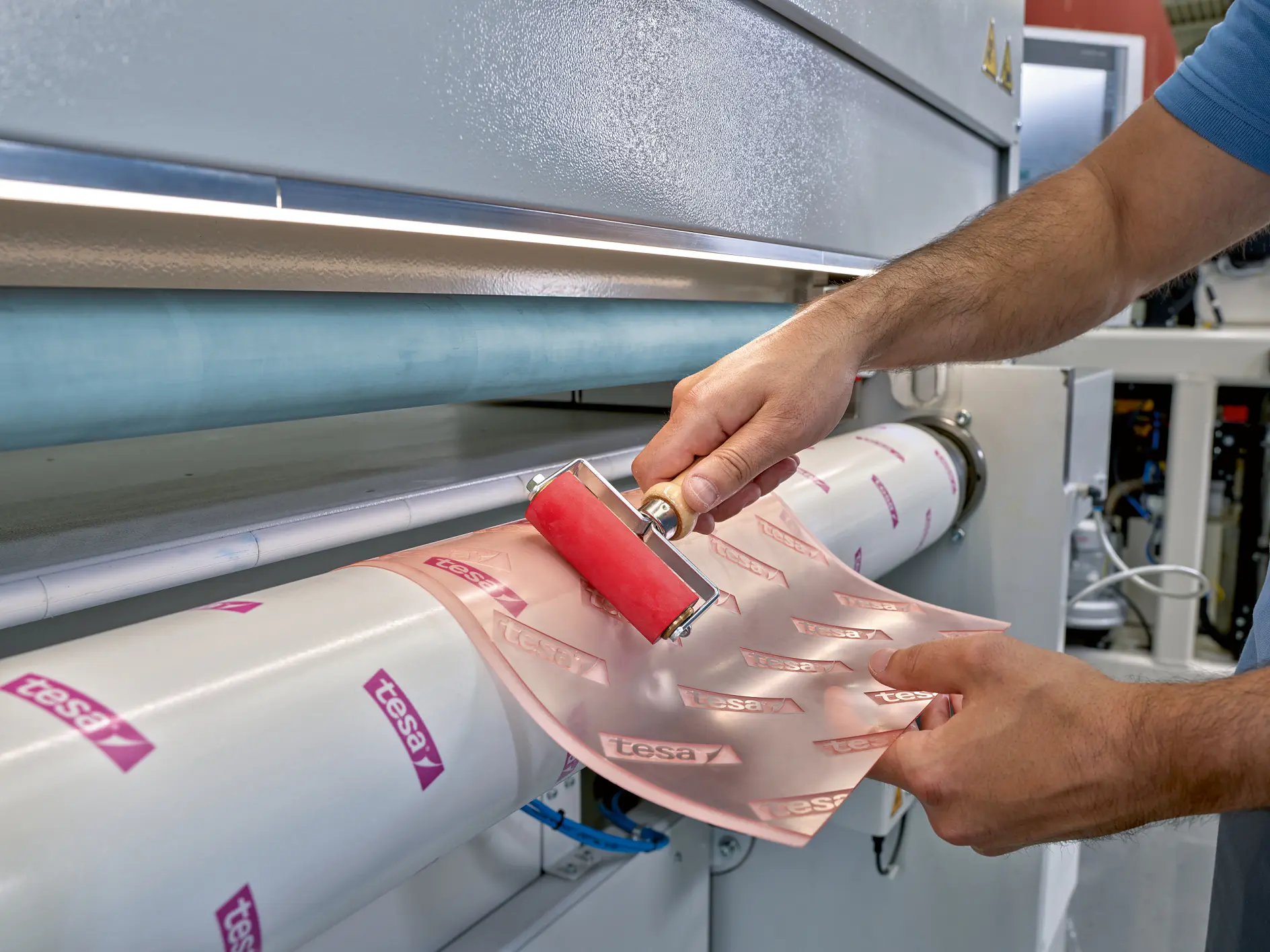 A person uses a red roller to smooth pink adhesive paper labeled tesa tape onto a cylindrical surface, part of a large industrial machine. (This text has been generated by AI)