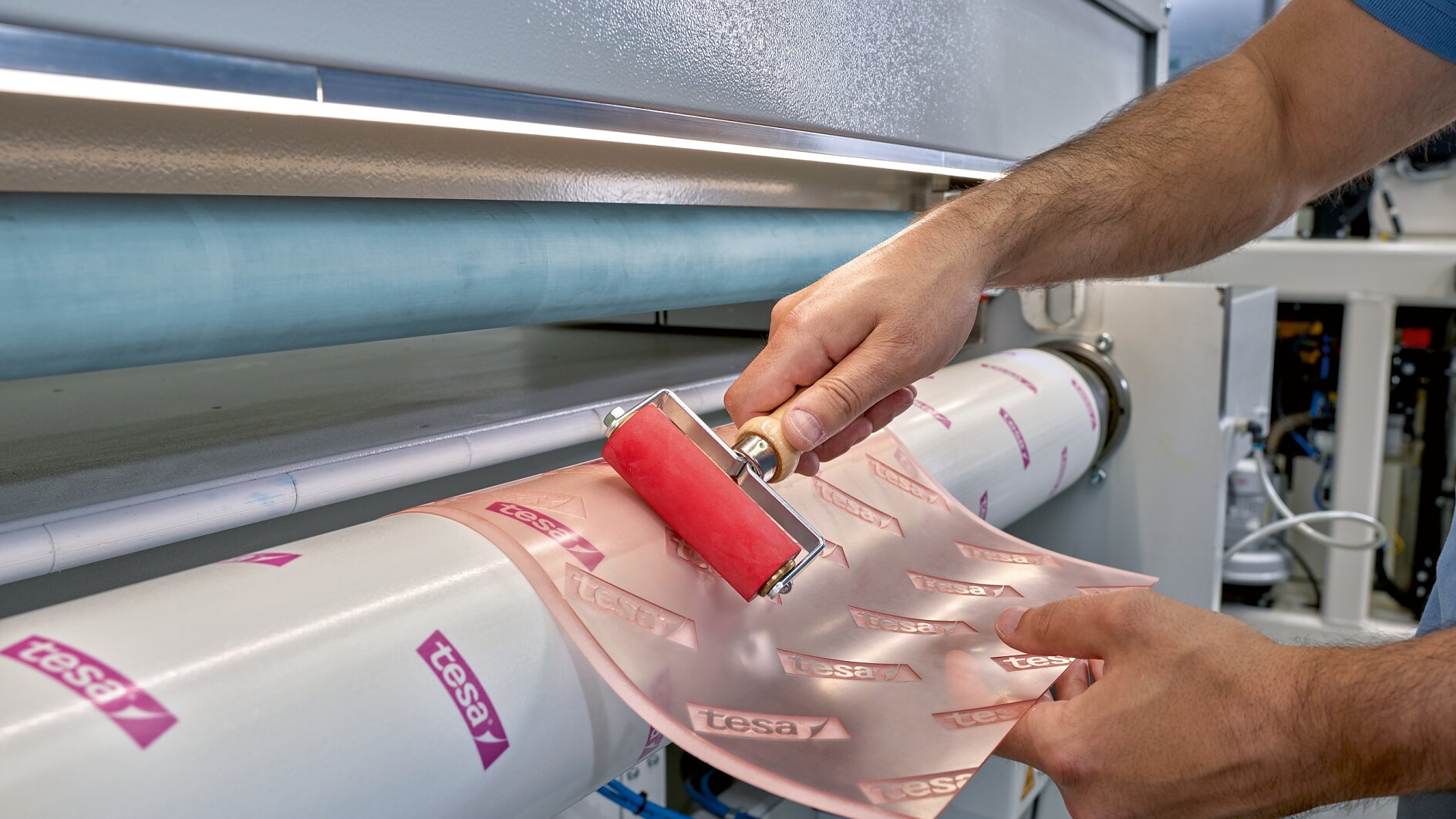 A person uses a red roller to smooth pink adhesive paper labeled tesa tape onto a cylindrical surface, part of a large industrial machine. (This text has been generated by AI)