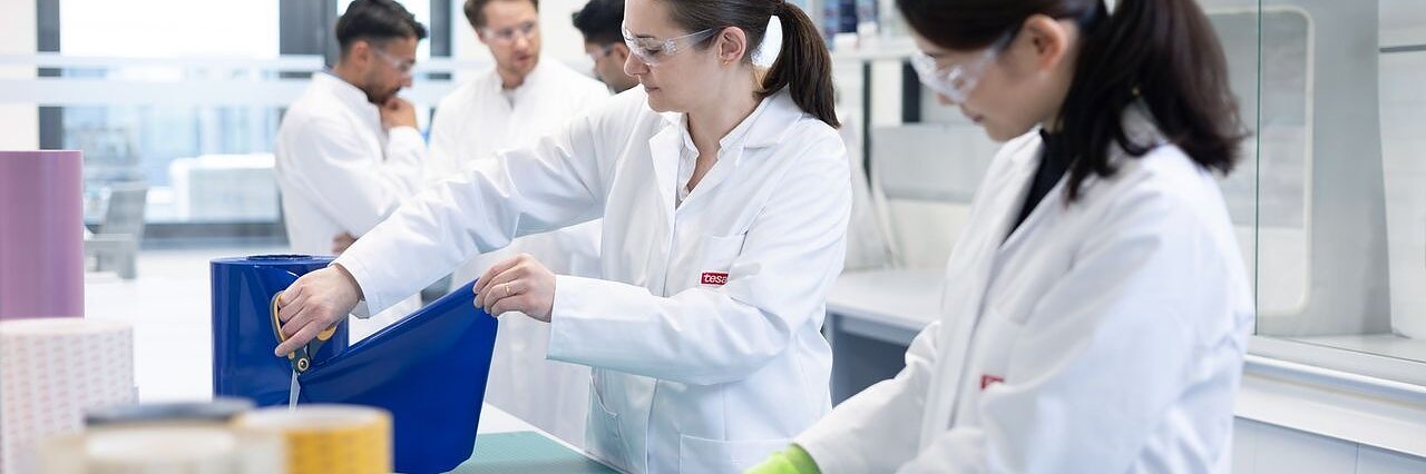 Two scientists in lab coats and safety glasses work with large rolls of tesa tape, while others talk nearby amid lab shelves and equipment.