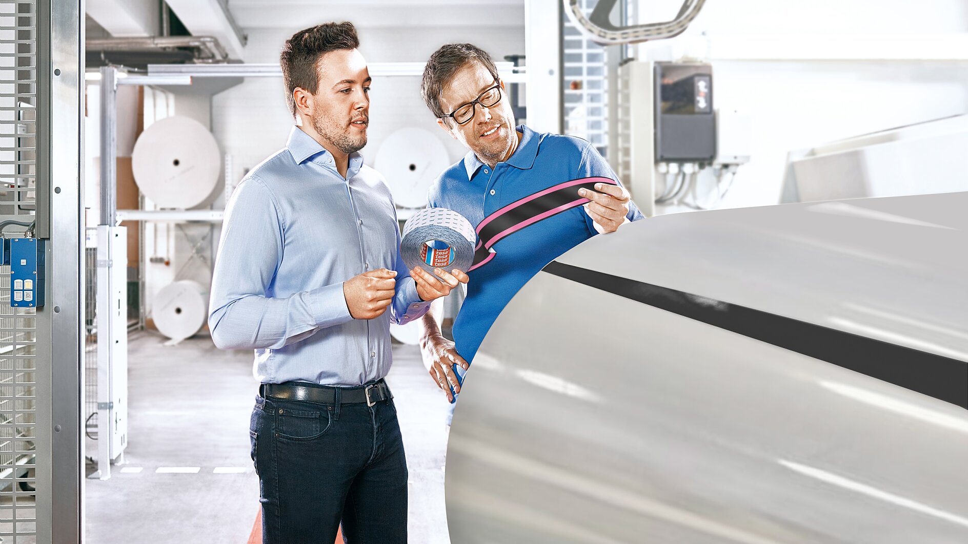 Two men in a manufacturing facility are inspecting a large industrial roller. One holds a roll of tesa tape and points to it with a tool. Coiled materials and machinery are visible in the background, suggesting a production environment. (This text has been generated by AI)