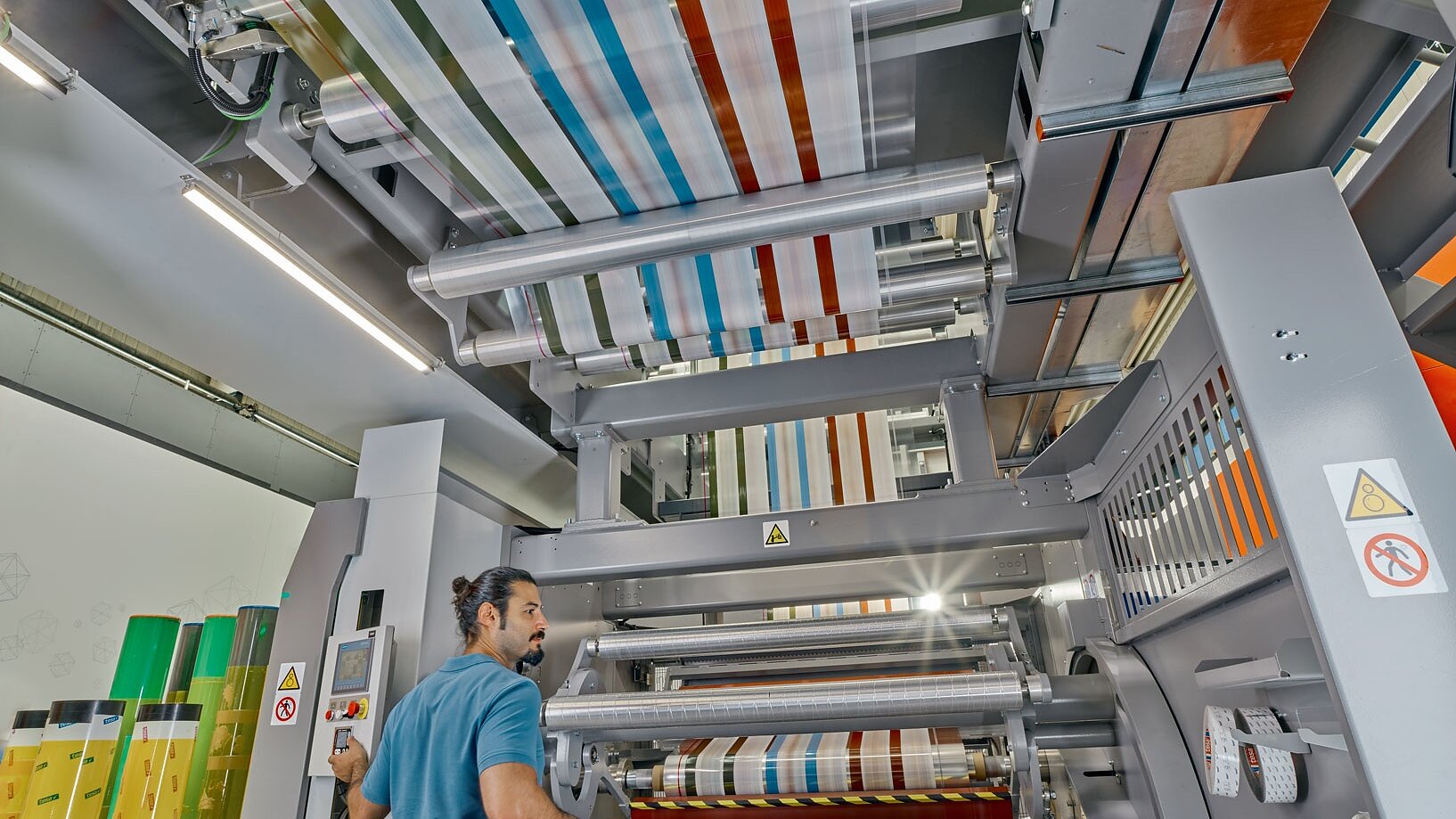 A man operates a large industrial printing machine with multiple rollers, producing colorful striped material. He uses tesa tape for securing certain parts of the process. The machine is surrounded by various safety signs, and the area is well-lit with overhead lights. (This text has been generated by AI)
