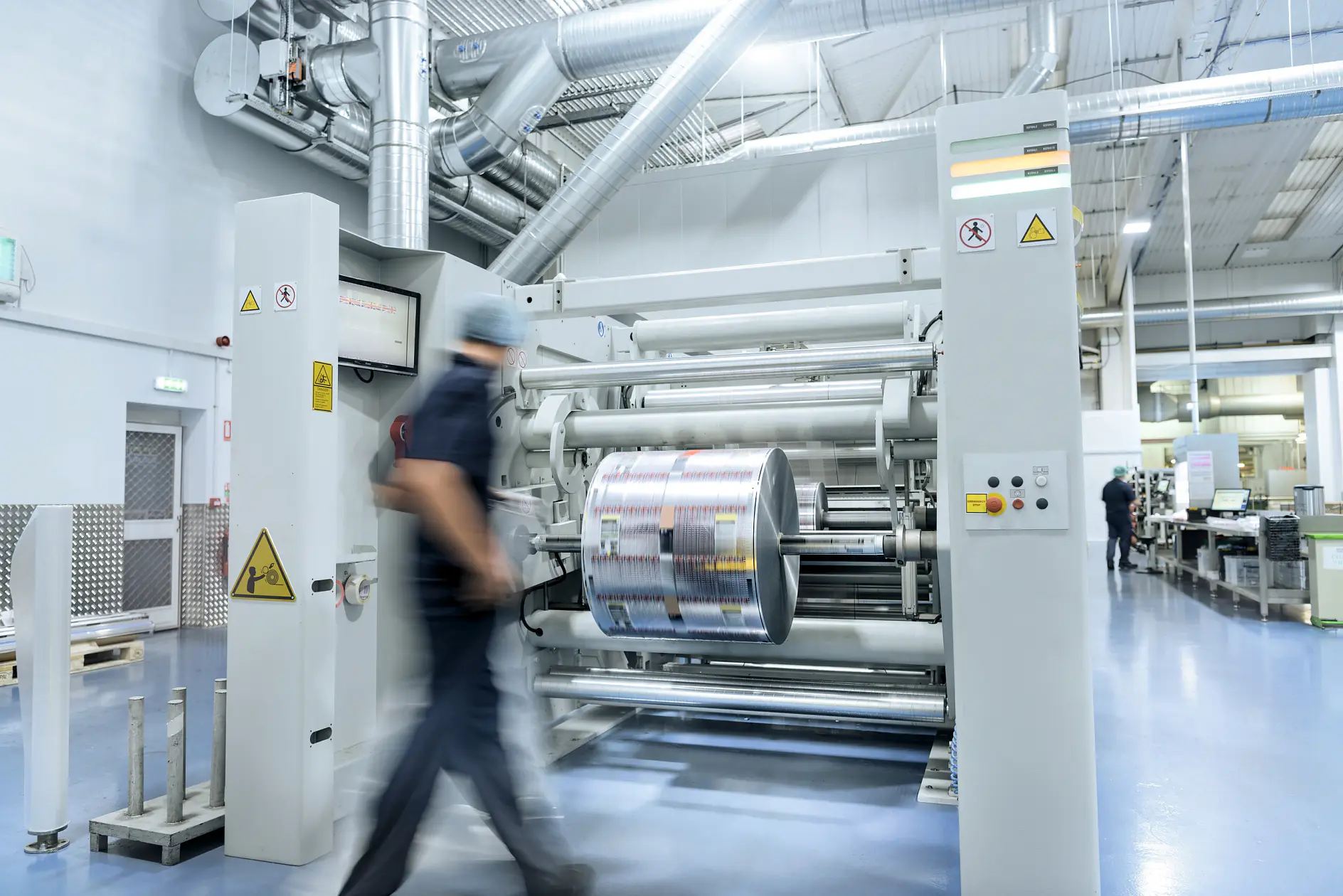 worker in navy uniform walking past large industrial printing machine in bright, spacious factory setting with metal pipes and equipment (This text has been generated by AI)
