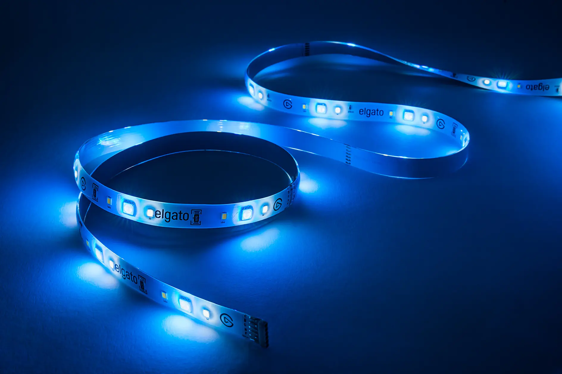 A flexible LED light strip emits a bright blue glow against a dark background. The strip is coiled in a loose, S-shaped pattern, showcasing individual LED components and tesa tape markings on the surface. (This text has been generated by AI)