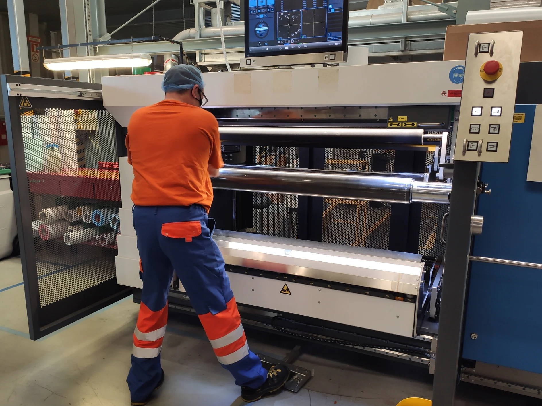 A worker in an orange shirt and blue pants operates a large industrial machine with rollers in a factory setting. He is facing the machine, which has a control panel at the top. Various materials, including tesa tape, are visible in the background. (This text has been generated by AI)
