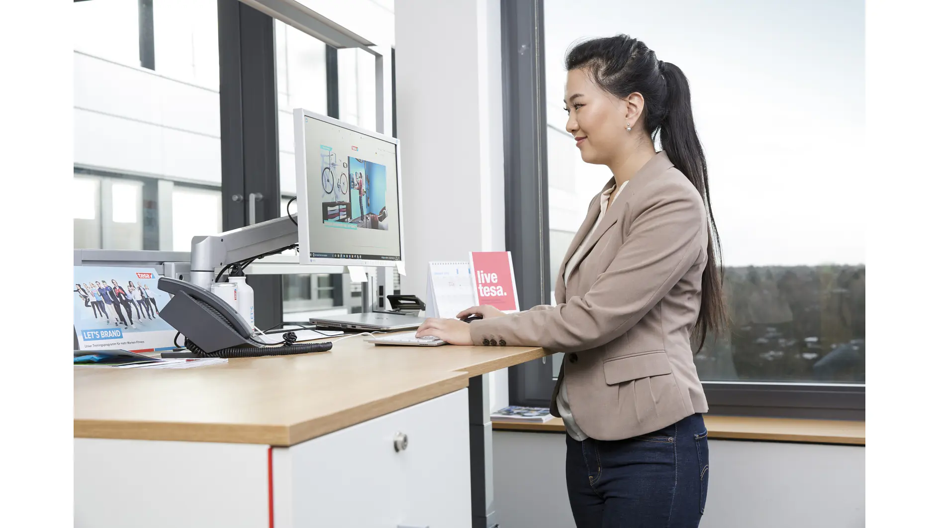 A woman in a beige blazer works at a standing desk with a computer monitor. The monitor displays a promotional webpage. She is in a modern office with large windows. A sign on the desk reads live tesa tape. (This text has been generated by AI)