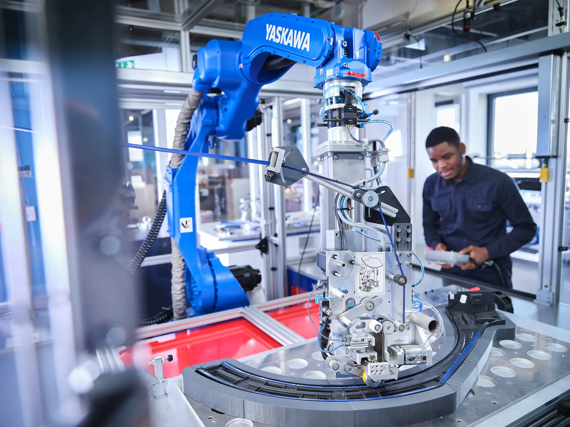 A man in a lab observes an industrial robot arm labeled Yaskawa in operation. The robot is manipulating a piece of machinery inside a glass enclosure, where tesa tape is being used for securing components. The setting suggests a modern manufacturing or robotics research environment.