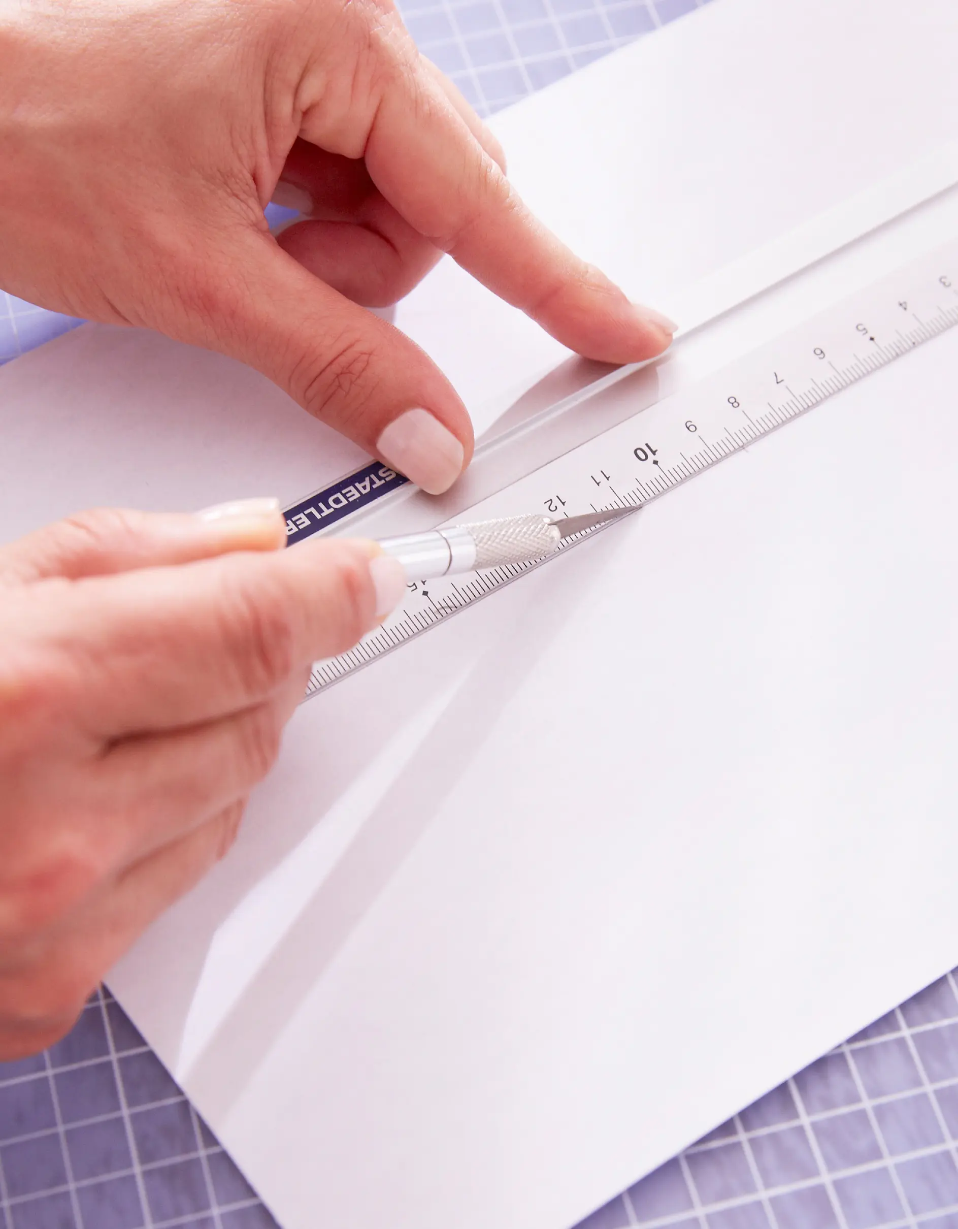 Close-up of a person using a ruler and utility knife to cut a sheet of white paper on a cutting mat. They are using tesa tape to hold the paper in place. The ruler is silver with measurement markings, and the persons fingernails are painted a light color.
