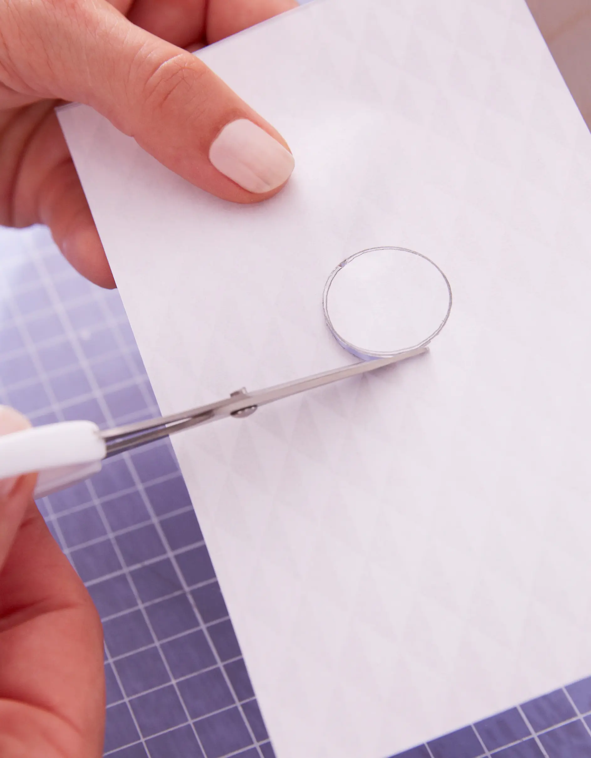 A hand with white nail polish holds a piece of patterned white paper. Another hand uses scissors to cut out a circular shape from the paper, secured with tesa tape. A blue grid cutting mat is visible underneath.